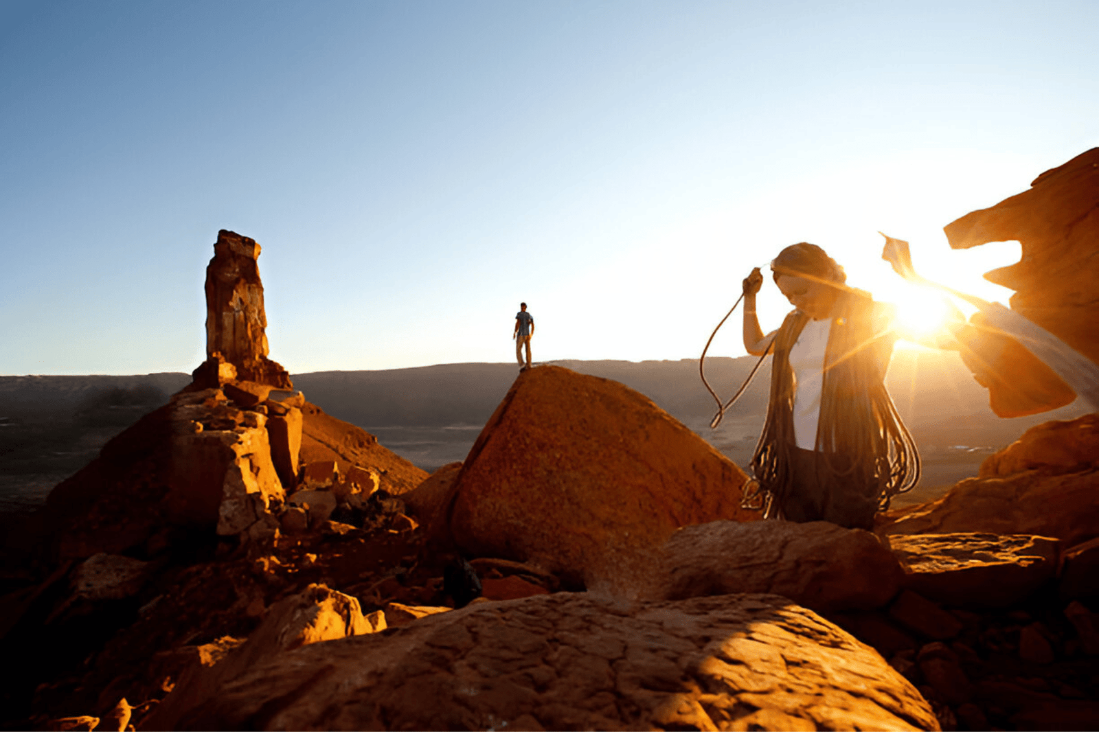 A happy young female rock climber coils her climbing rope at sunset as her husband stands on a rock in the distance looking for the trail back to camp.